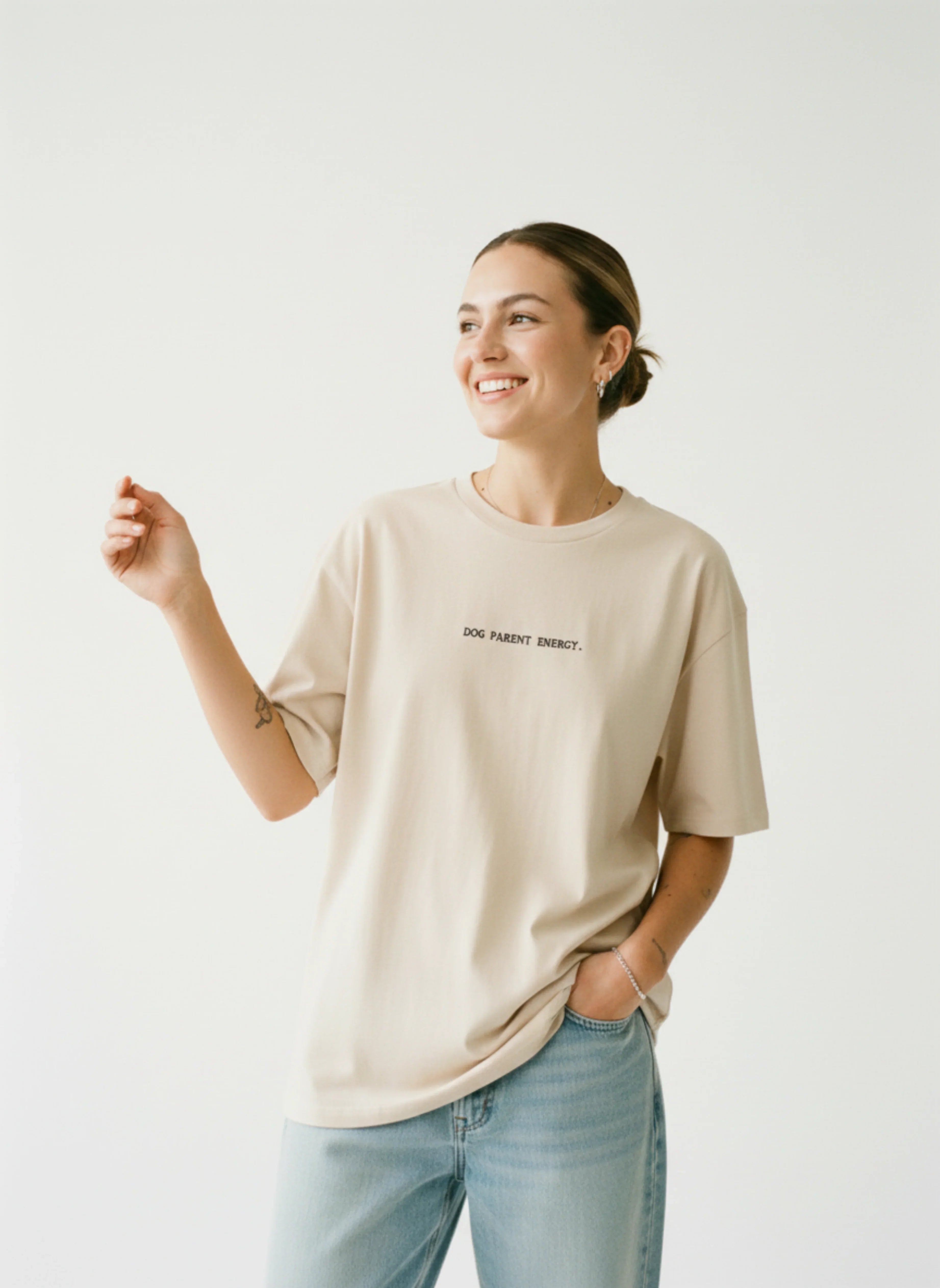 Woman wearing beige 'Dog Parent Energy' tee with jeans, smiling against white background